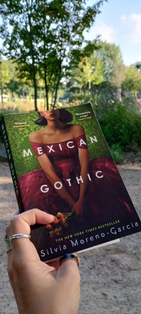 Foto von einer Hand, die den Roman "Mexican Gothic" hält. Auf dem Cover des Buchs zu sehen ist eine junge Frau in einem roten, schulterlosen Kleid, die einen Strauss gold-gelber Blumen in den Händen hält. Ihr Gesicht ist ab den Augen aufwärts aufgrund des Bildauschnitts nicht erkennbar.